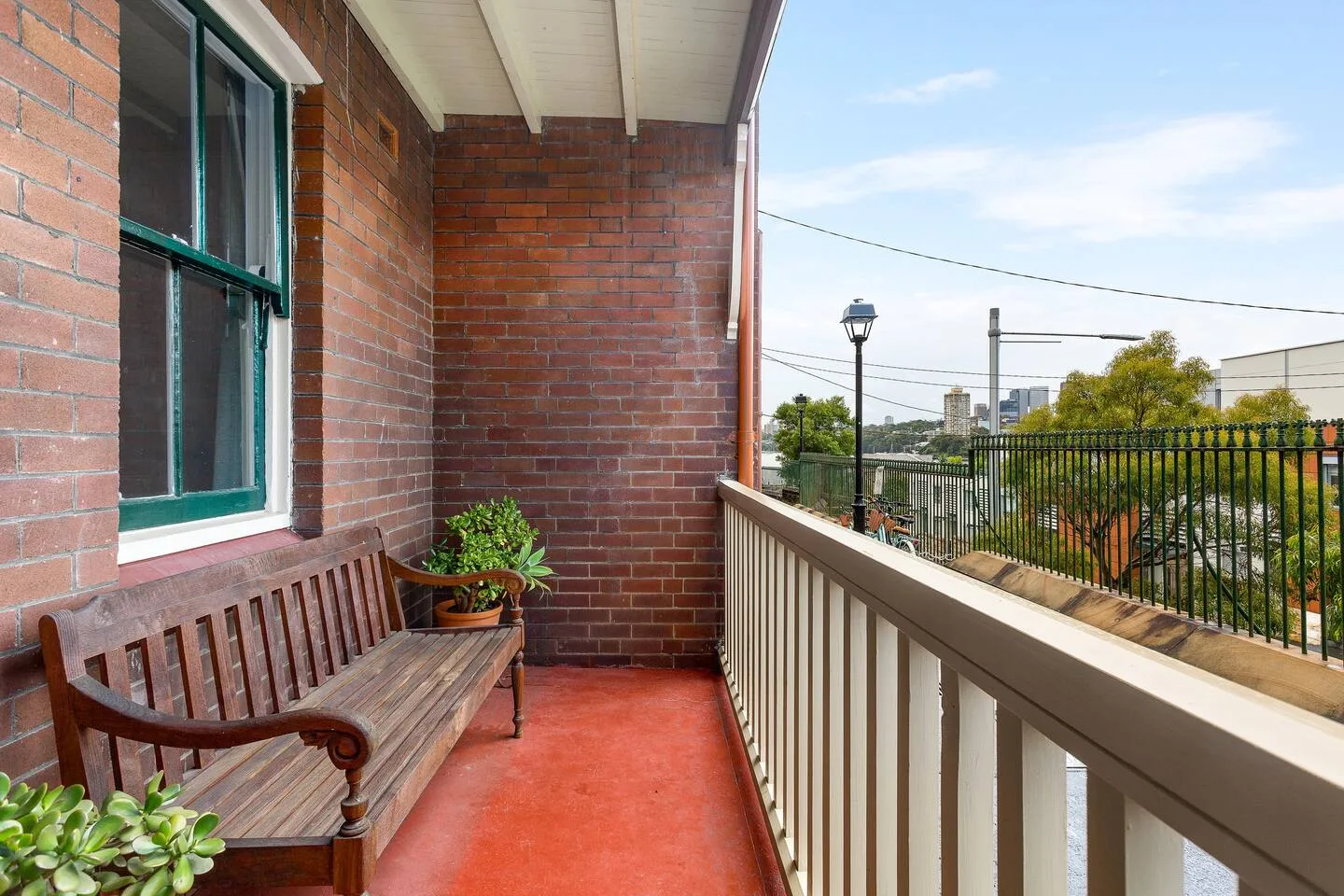 A covered balcony with a wooden bench and railing overlooks trees and nearby buildings.