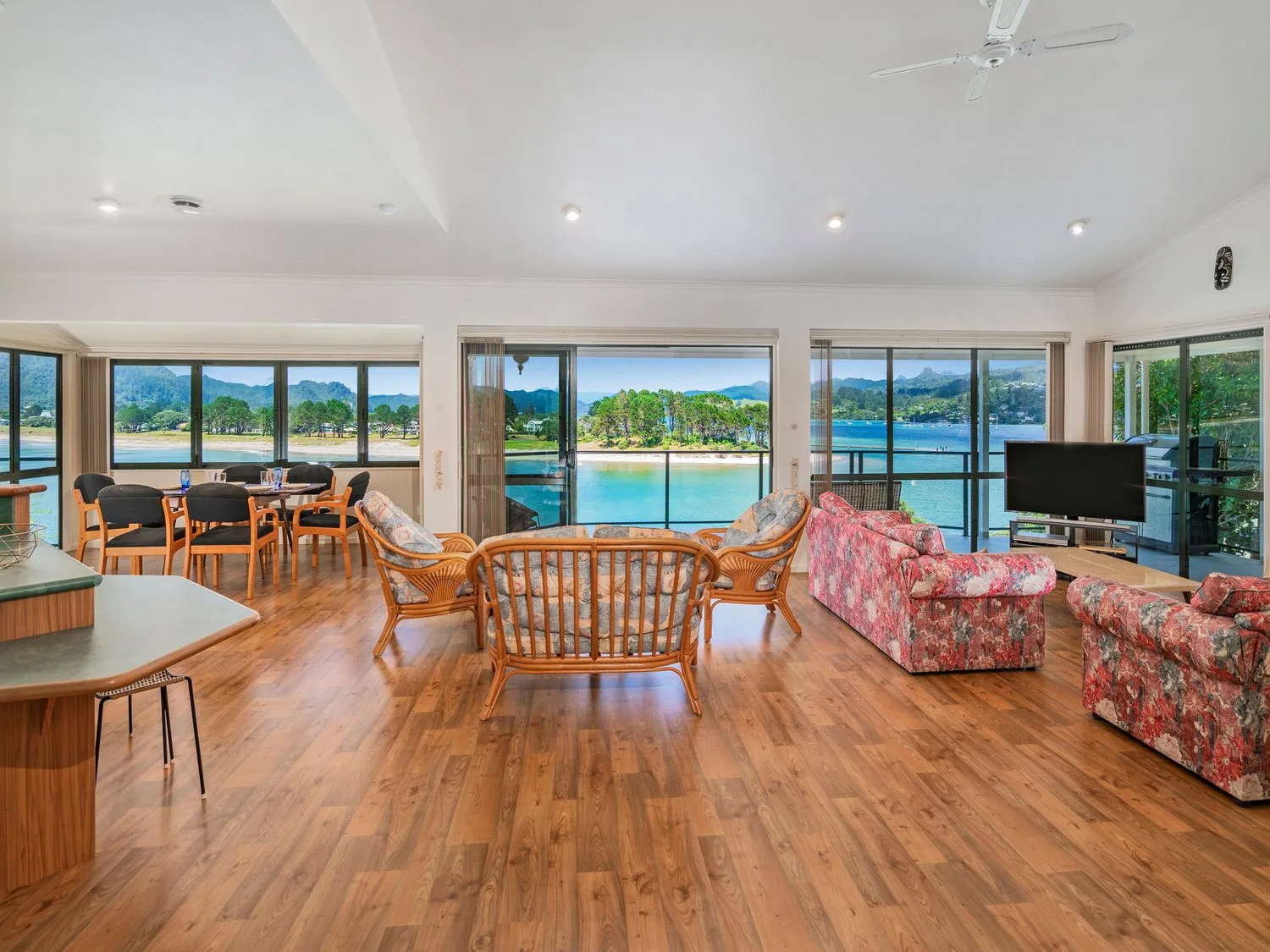 Una sala de estar con muebles de estampado floral y de mimbre y una mesa de comedor, con vistas al agua y las montañas en Pohutukawa on Paku - Tairua Holiday Home, en Tairua.