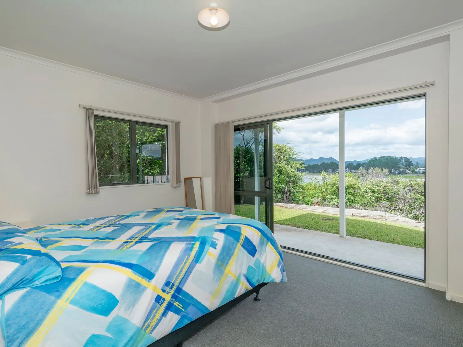 Un dormitorio con una cama cubierta con ropa de cama azul estampada y grandes puertas correderas de vidrio que dejan ver un jardín y vistas a las montañas en Pohutukawa on Paku - Tairua Holiday Home, en Tairua.