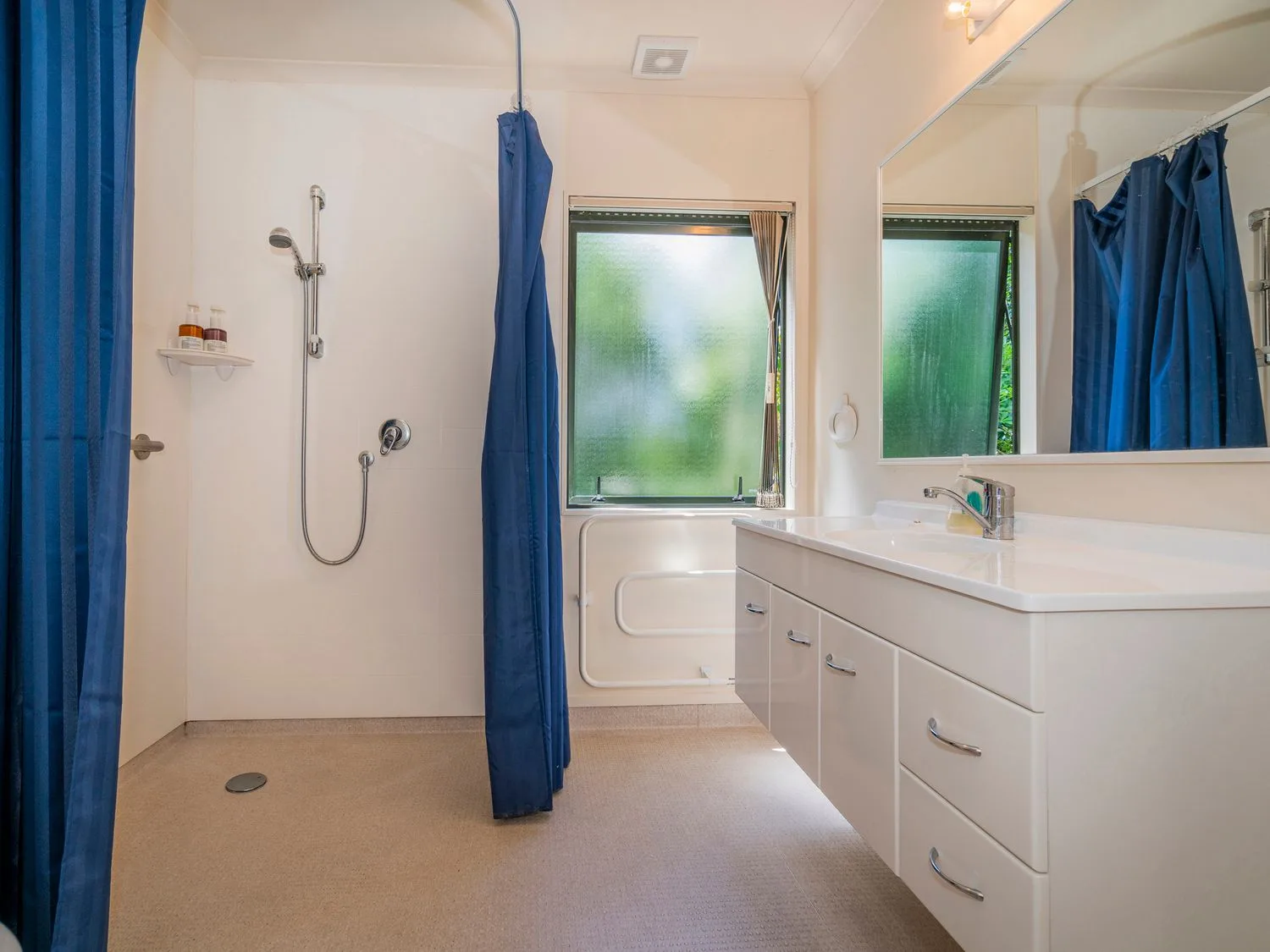 Un baño con una zona de ducha con cortinas azules, un lavabo con armarios de almacenamiento y una ventana de vidrio esmerilado en Pohutukawa on Paku - Tairua Holiday Home, en Tairua.