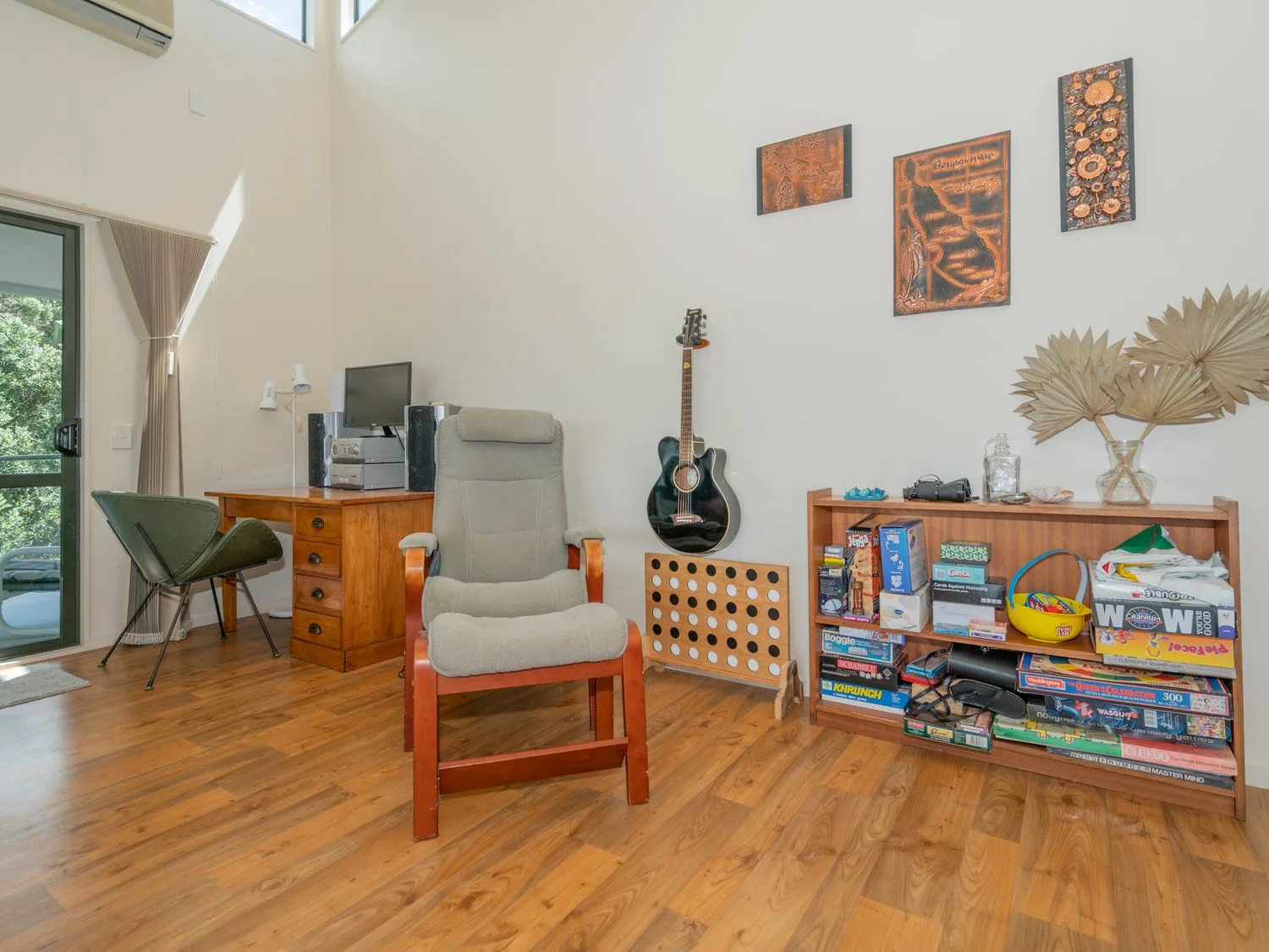 Una habitación con un escritorio y una silla de madera, un sillón acolchado, una guitarra colgada en la pared, una estantería de madera con juegos de mesa y un jarrón de vidrio con hojas secas en Pohutukawa on Paku - Tairua Holiday Home, en Tairua.
