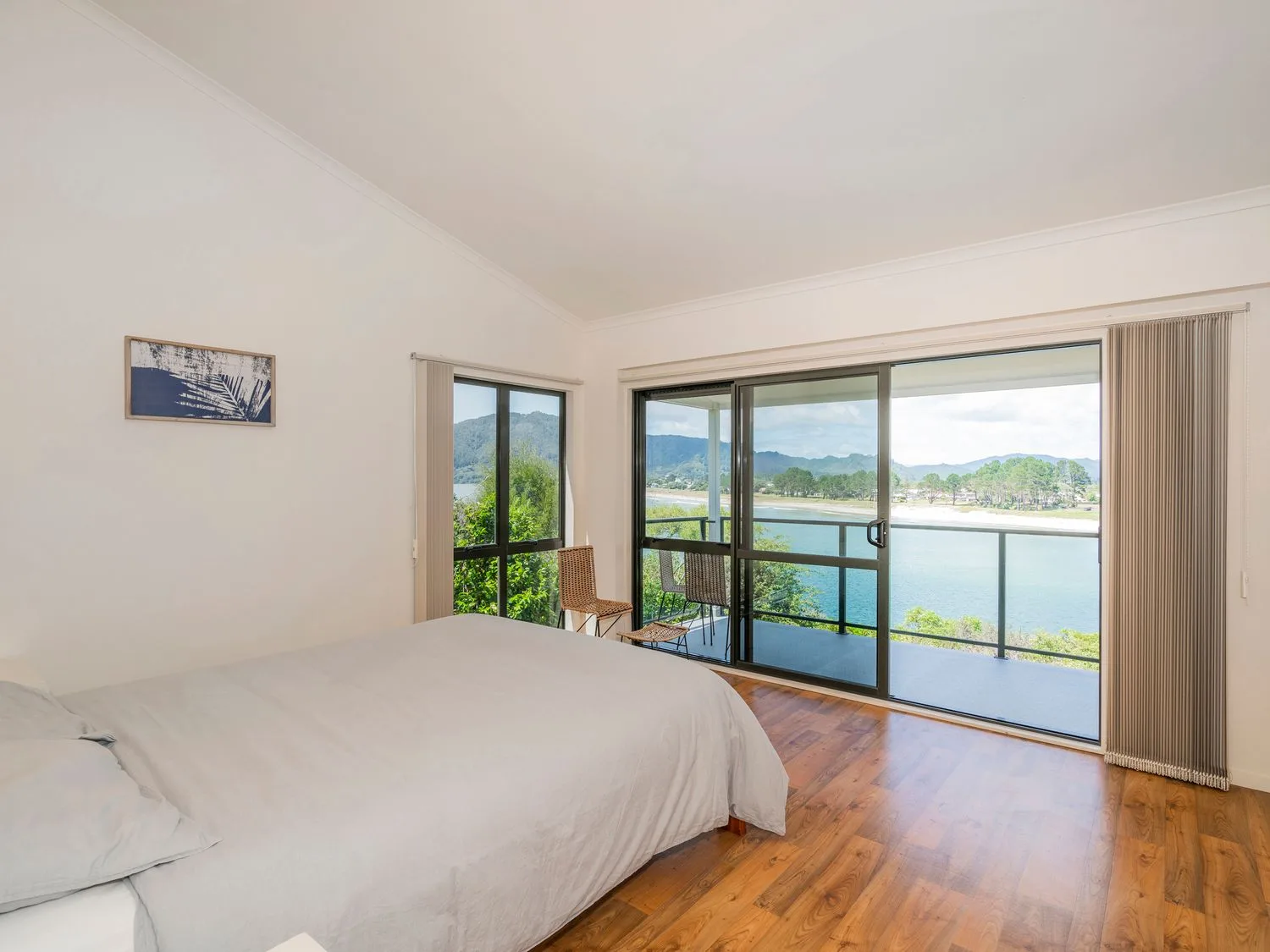 Un dormitorio con una cama y dos sillas junto a puertas correderas de vidrio con vistas a una masa de agua y colinas en Pohutukawa on Paku - Tairua Holiday Home, en Tairua.