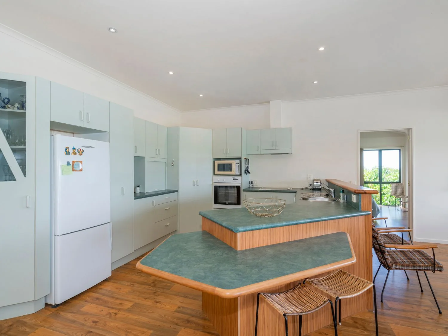 Una cocina con encimeras verdes, armarios blancos, un refrigerador blanco y taburetes en Pohutukawa on Paku - Tairua Holiday Home, en Tairua.