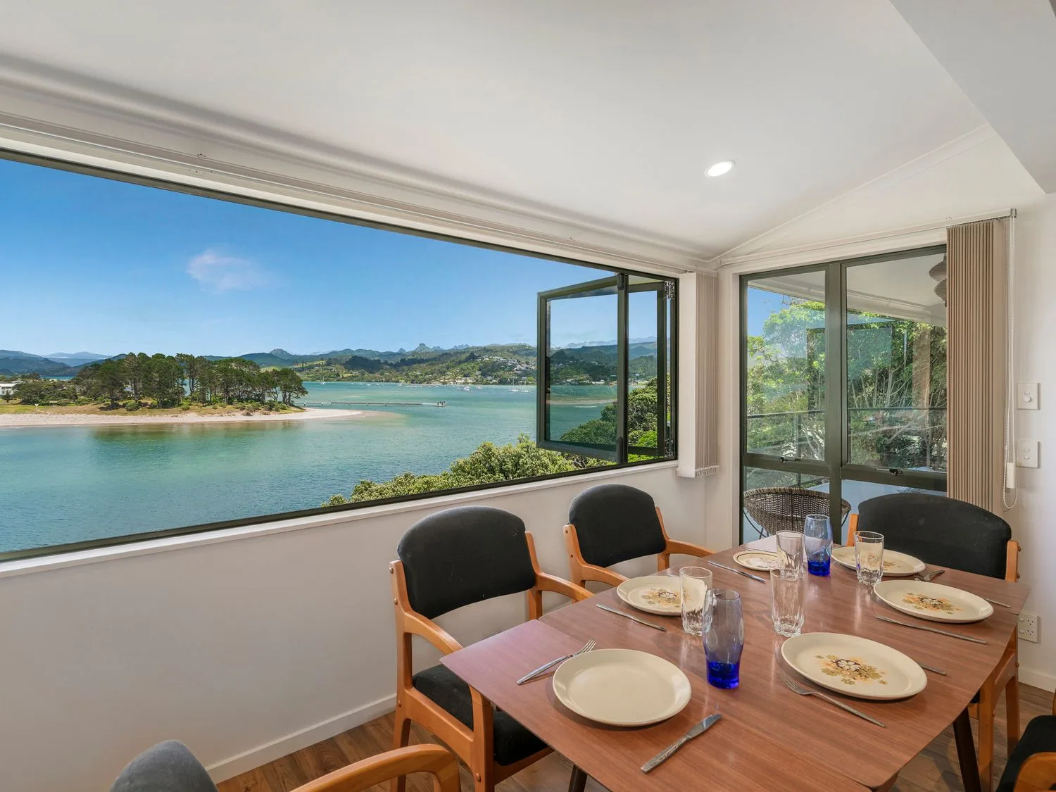 Un comedor con una mesa de madera puesta con platos y vasos junto a grandes ventanas con vistas a una masa de agua y árboles en Pohutukawa on Paku - Tairua Holiday Home, en Tairua.
