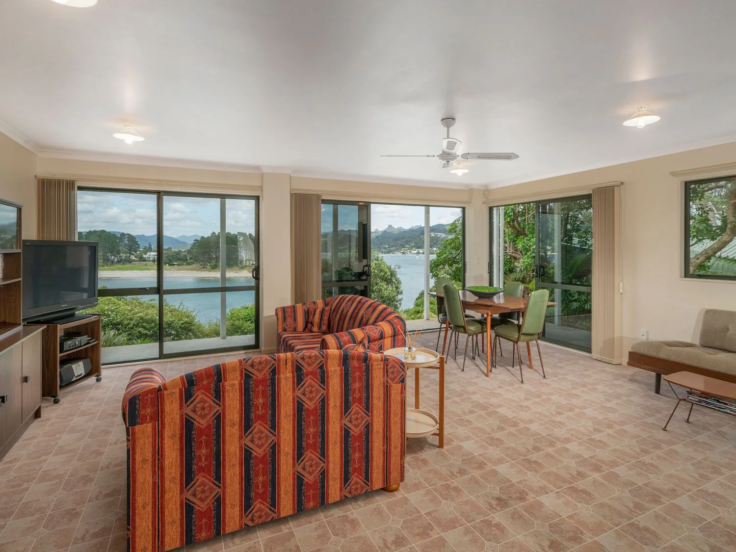 Una sala de estar con sofás estampados, una mesa de comedor con sillas y grandes ventanas con vistas a una masa de agua en Pohutukawa on Paku - Tairua Holiday Home, en Tairua.