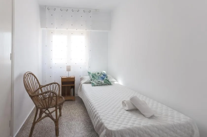 A bright bedroom with a single bed, bedside table, and wicker seating.