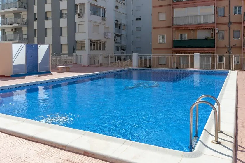 A blue swimming pool sits among apartment buildings, with a metal ladder at the edge.