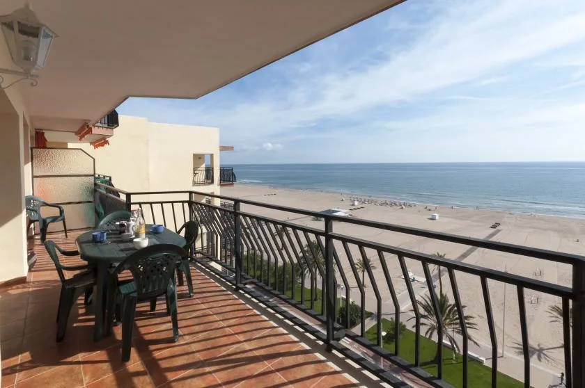 A furnished balcony overlooks a sandy beach and the ocean.