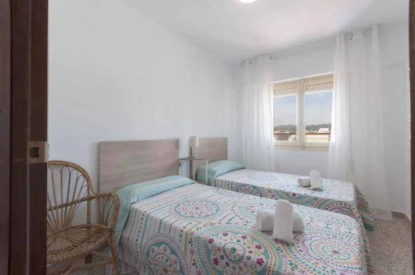 A bright bedroom with two beds, patterned bedding, and a window with sheer curtains.
