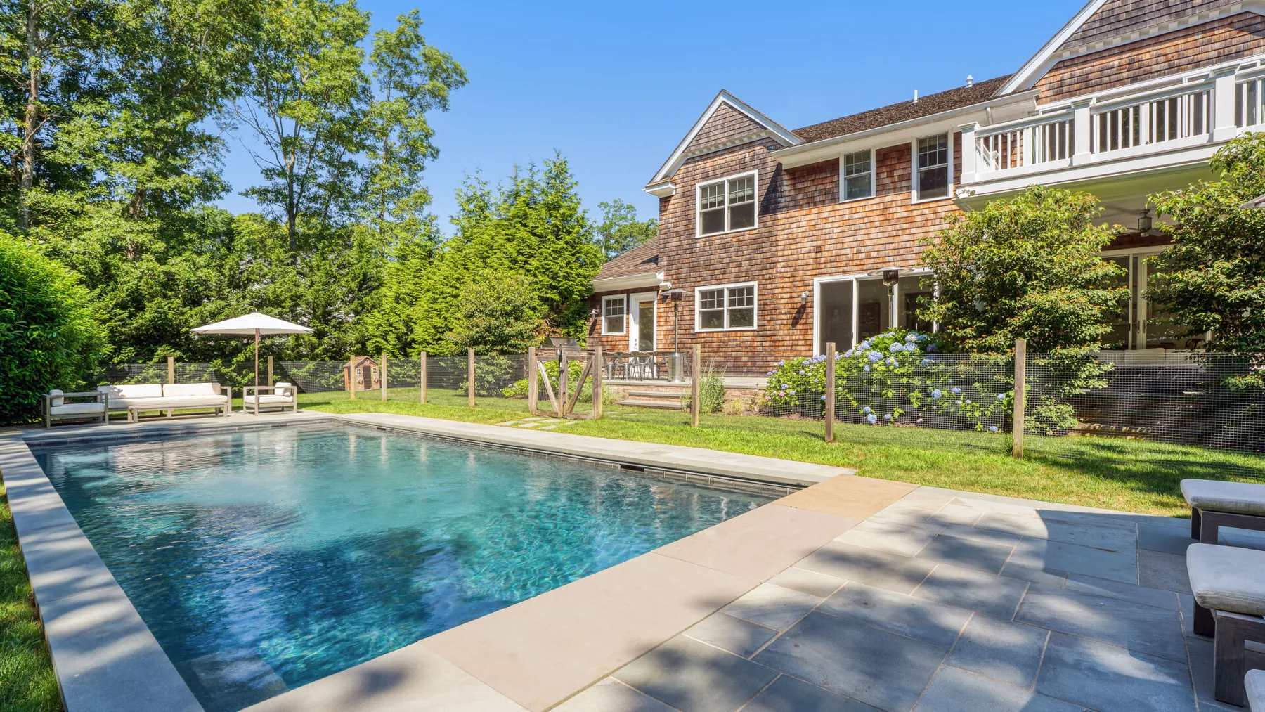 Una piscina exterior está junto a una casa grande y un patio pavimentado.