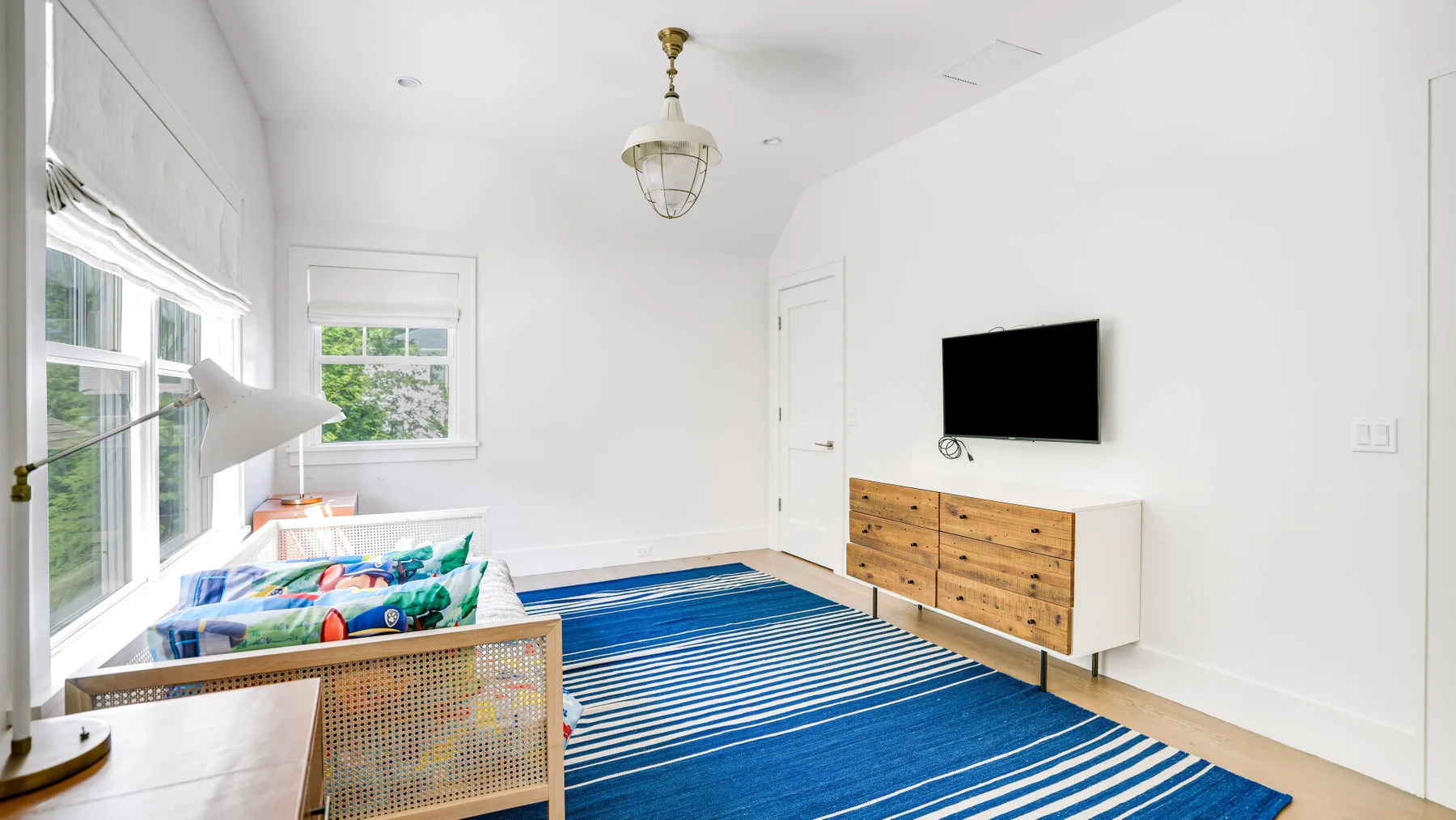 Un dormitorio luminoso con una cama, una alfombra azul a rayas y una cómoda bajo un televisor.