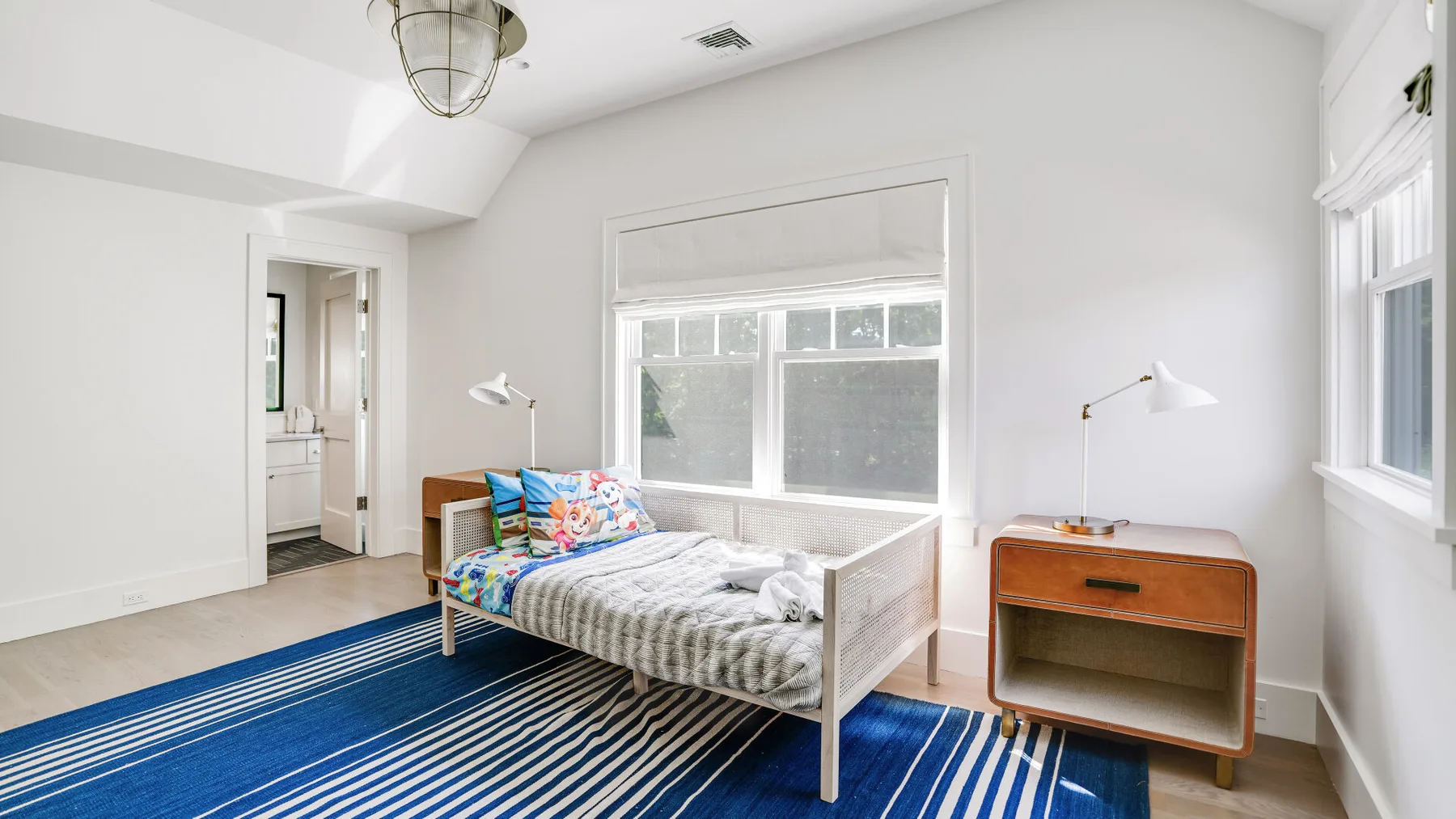Un dormitorio luminoso con una cama, una alfombra a rayas azules y ventanas.