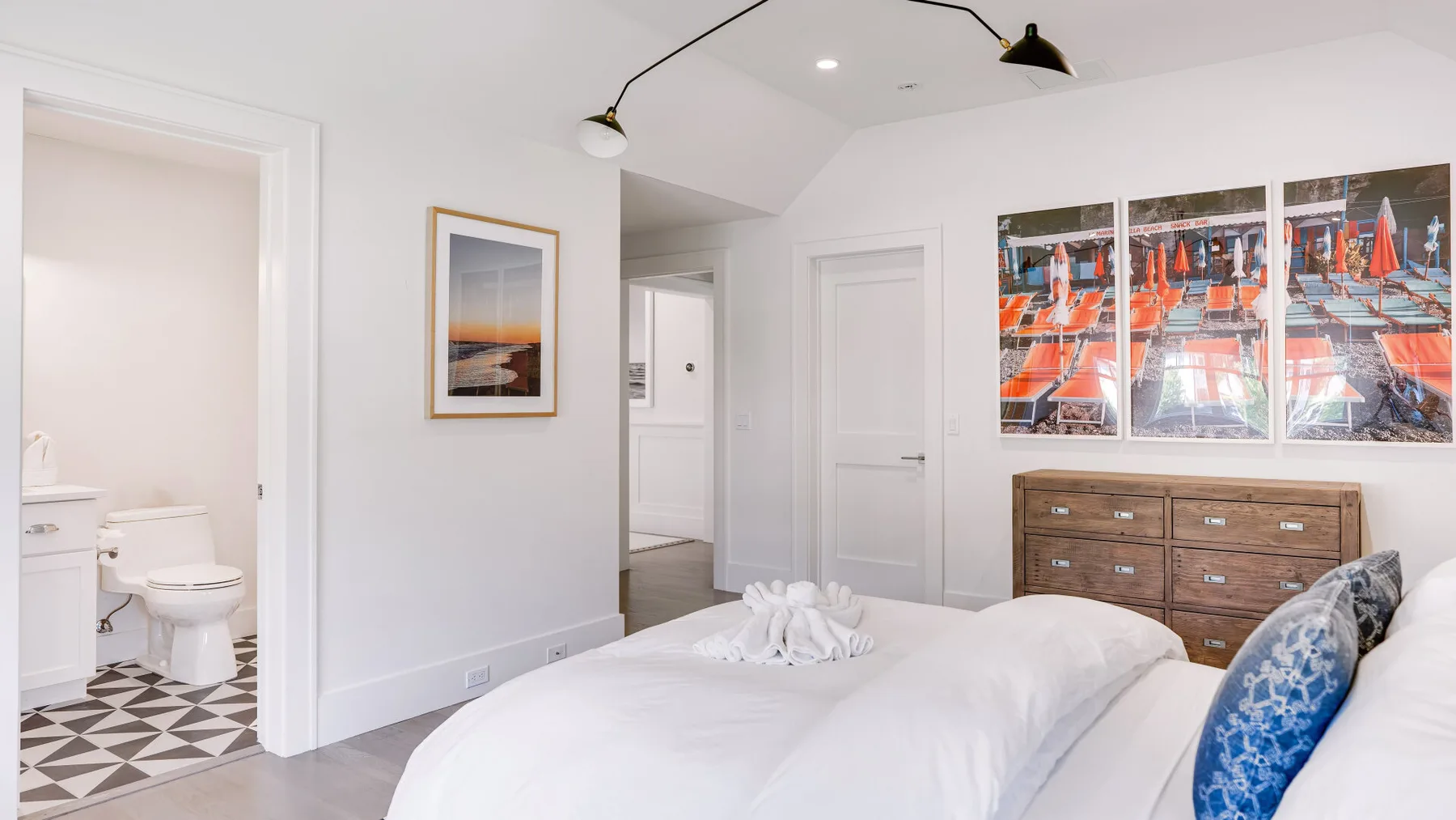 Un dormitorio luminoso con una cama blanca, una cómoda y arte colorido en la pared.