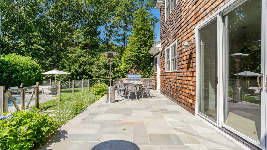 Un patio pavimentado junto a una casa con revestimiento de madera da a un jardín frondoso.