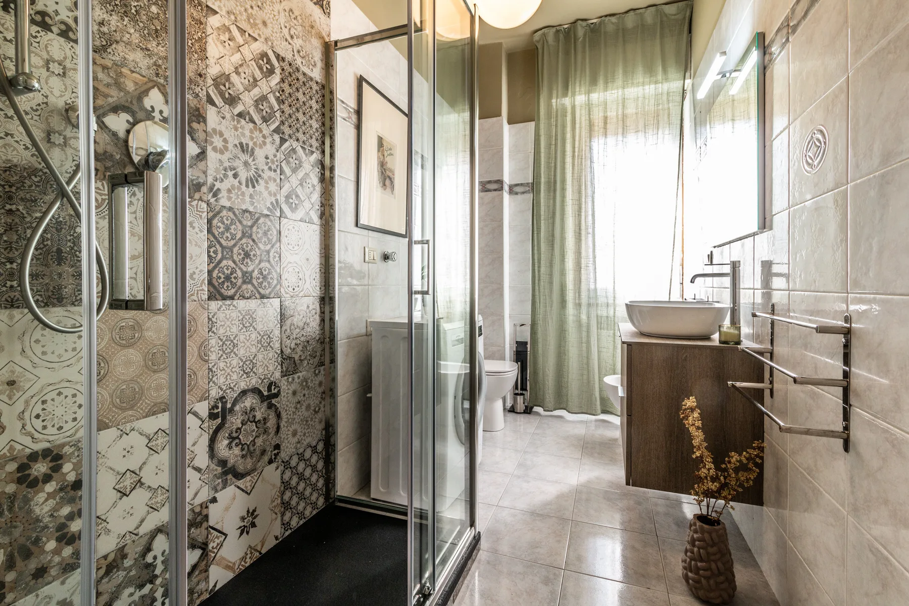 Un baño con ducha de vidrio, lavabo de recipiente y una ventana con cortina.