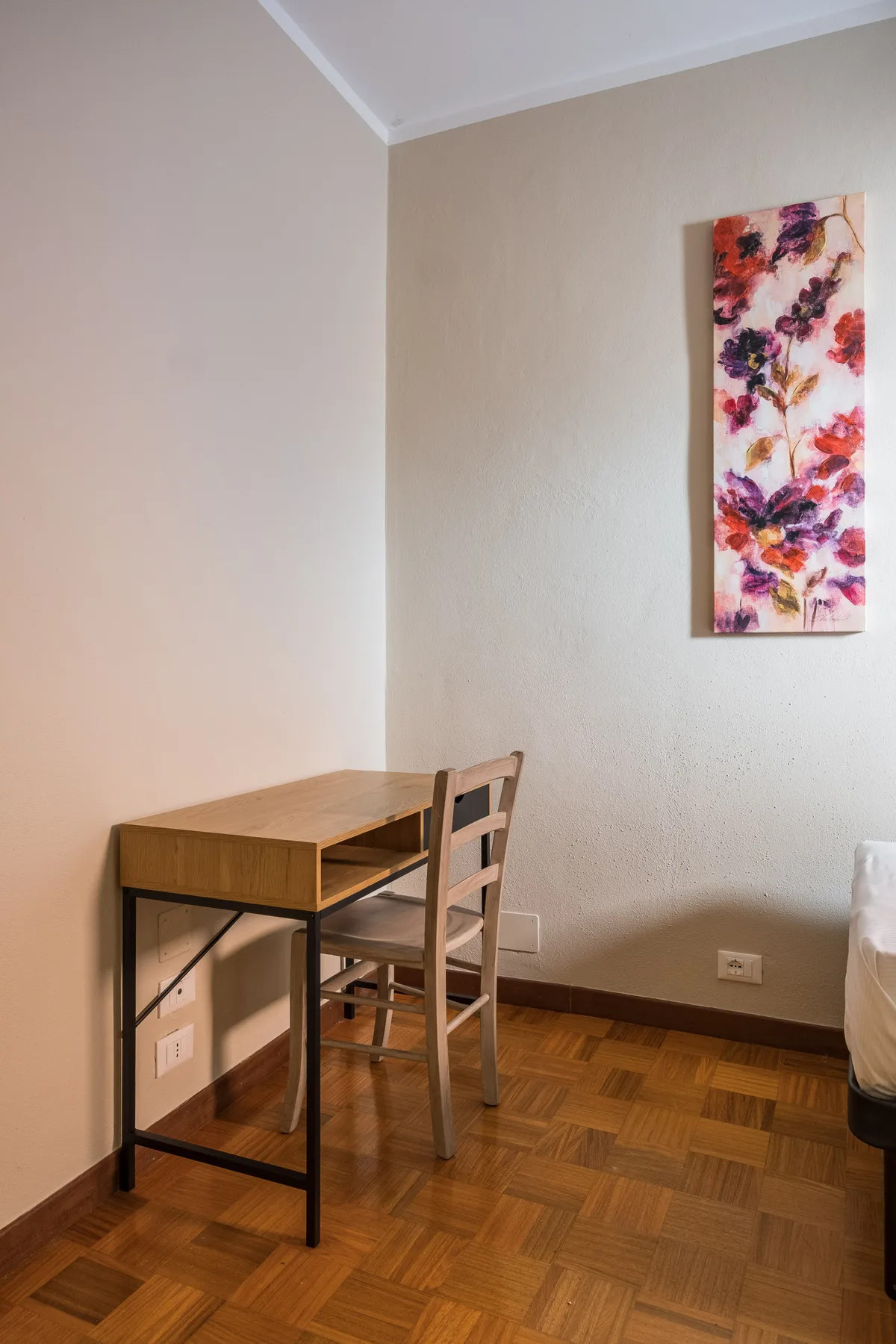 Un escritorio pequeño y una silla están bajo un cuadro floral en un dormitorio.