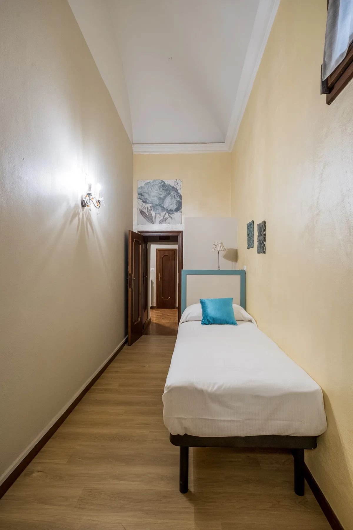 Un dormitorio estrecho tiene una cama individual con ropa de cama blanca y un cojín turquesa.