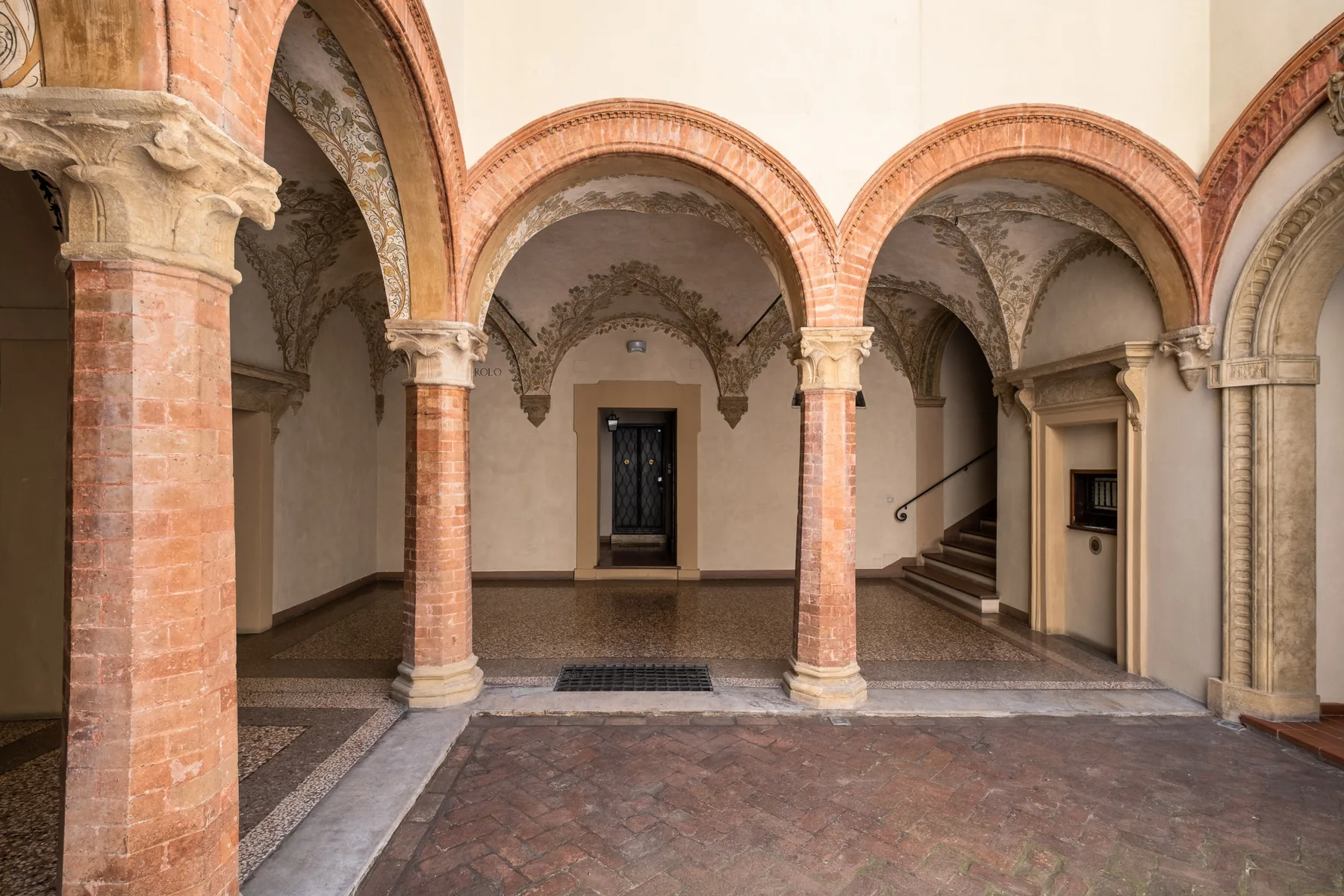 Un salón interior con arcos, columnas de ladrillo, suelo decorado y una escalera.