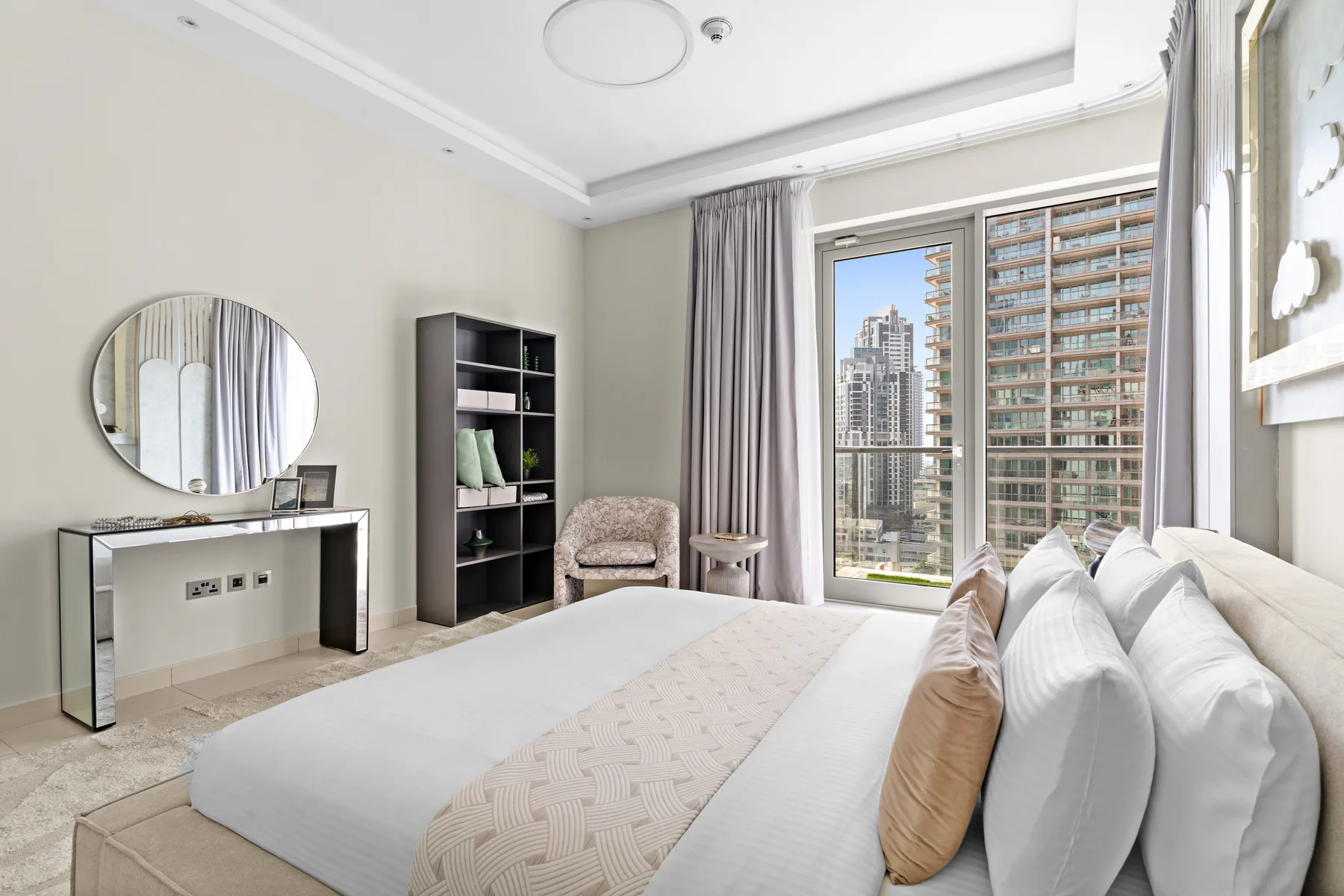 Un dormitorio luminoso con una cama grande, una consola y vistas de la ciudad por las ventanas.
