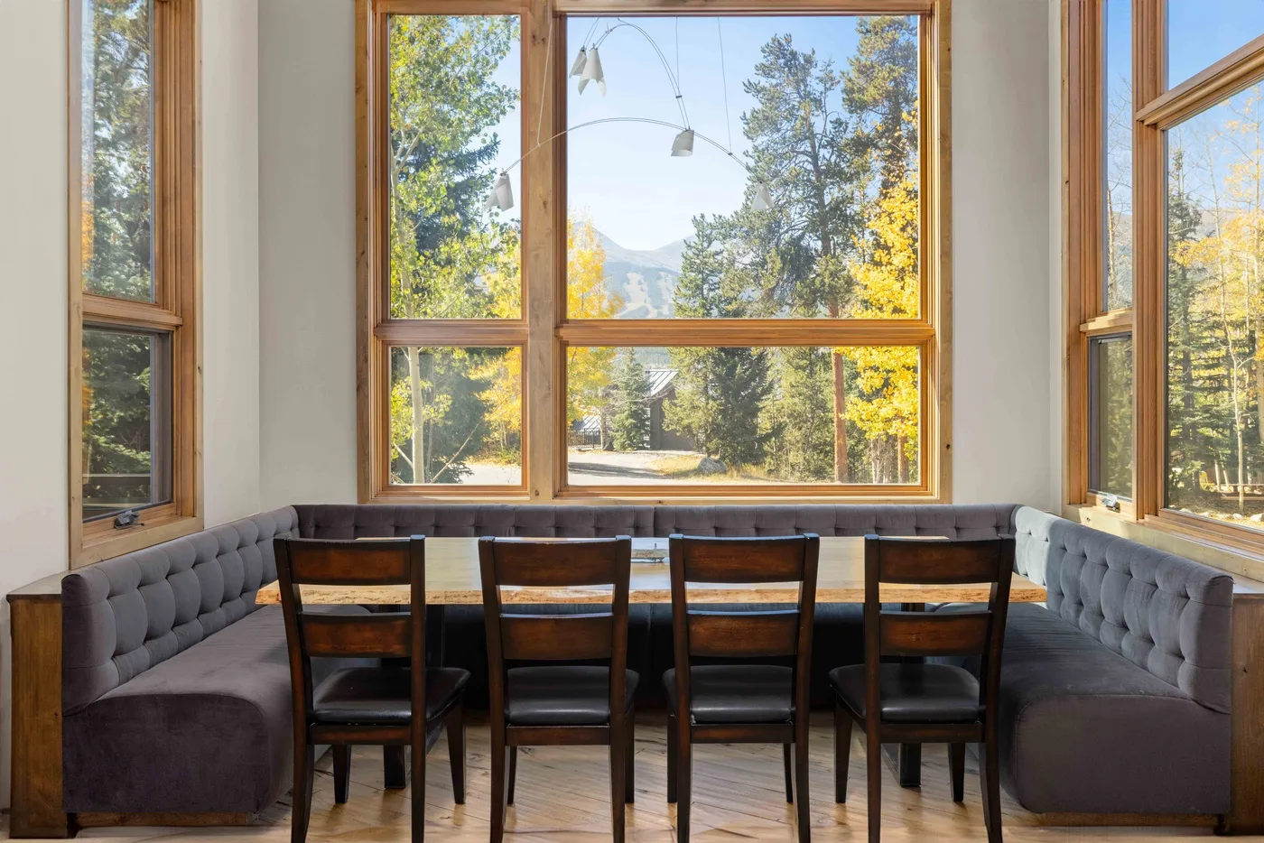 A dining area with a long table and seating beside large windows overlooking trees.