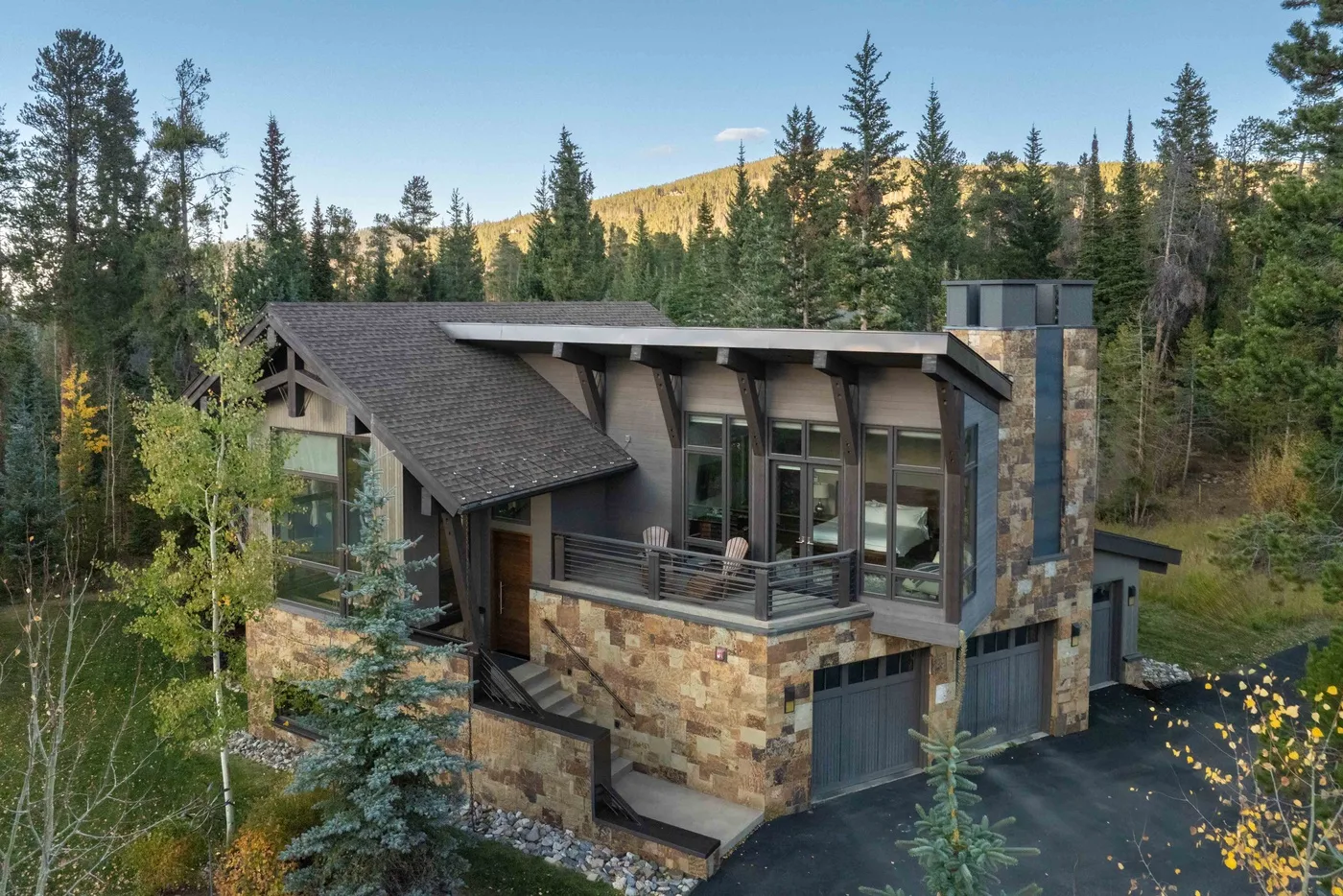 A modern house sits among evergreen trees, with a mountain ridge in the background.