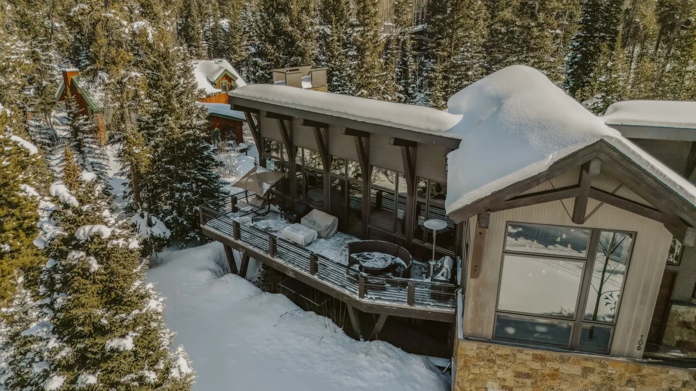 A snow-covered cabin sits among tall evergreen trees.