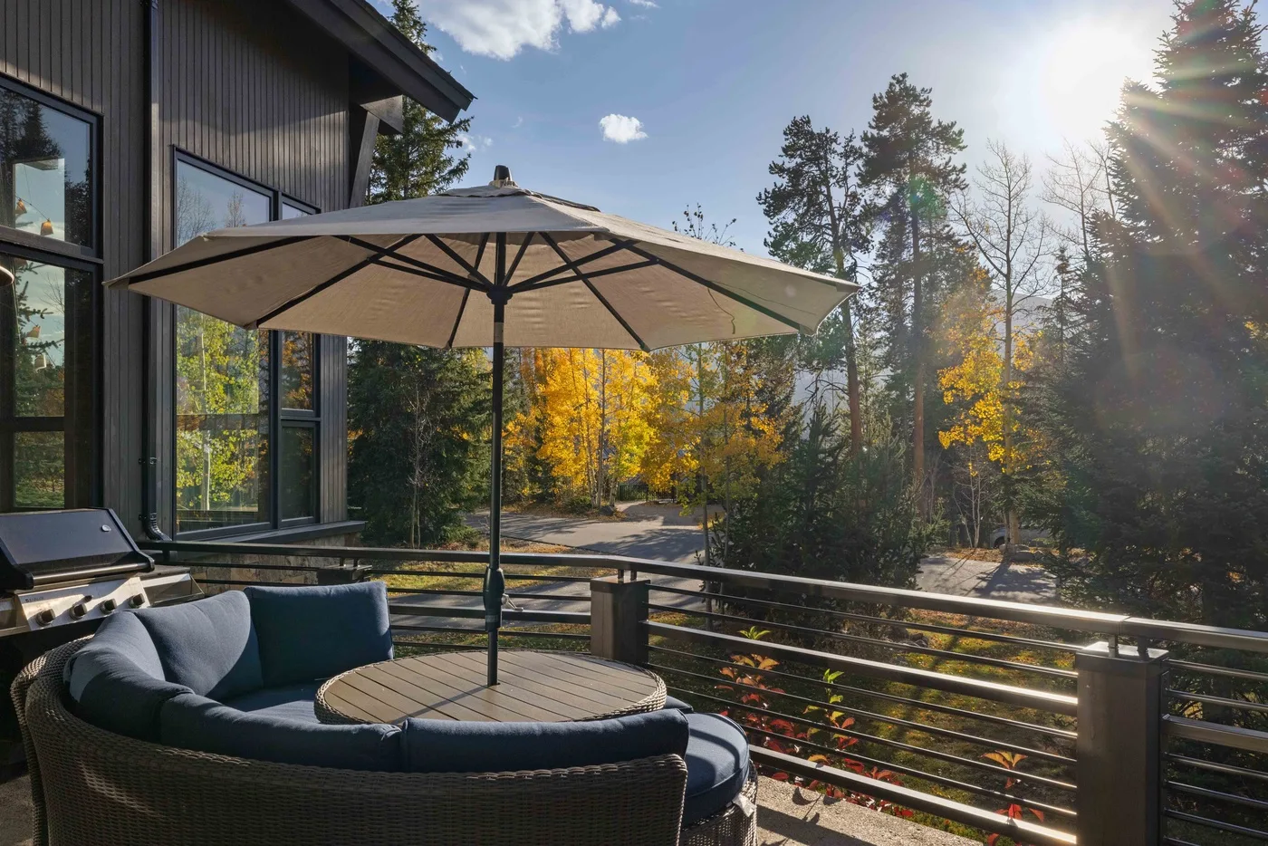 An outdoor deck with seating, a table, and an umbrella overlooks trees.