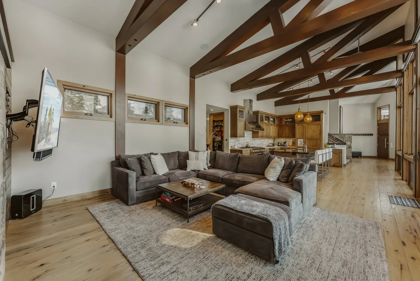 A living room with sectional seating and an open kitchen beneath exposed beams.
