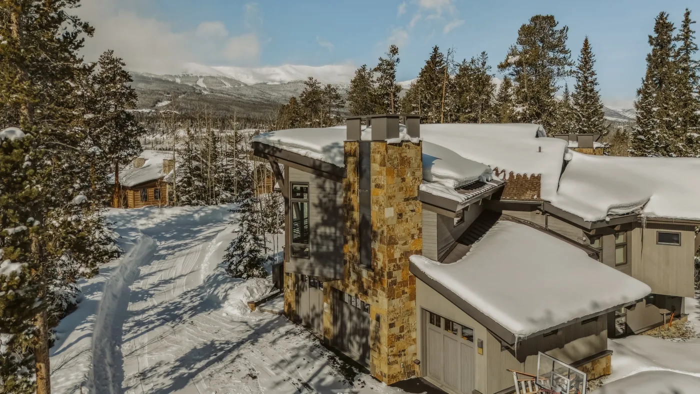 A snow-covered lodge sits among evergreen trees with mountains in the distance.