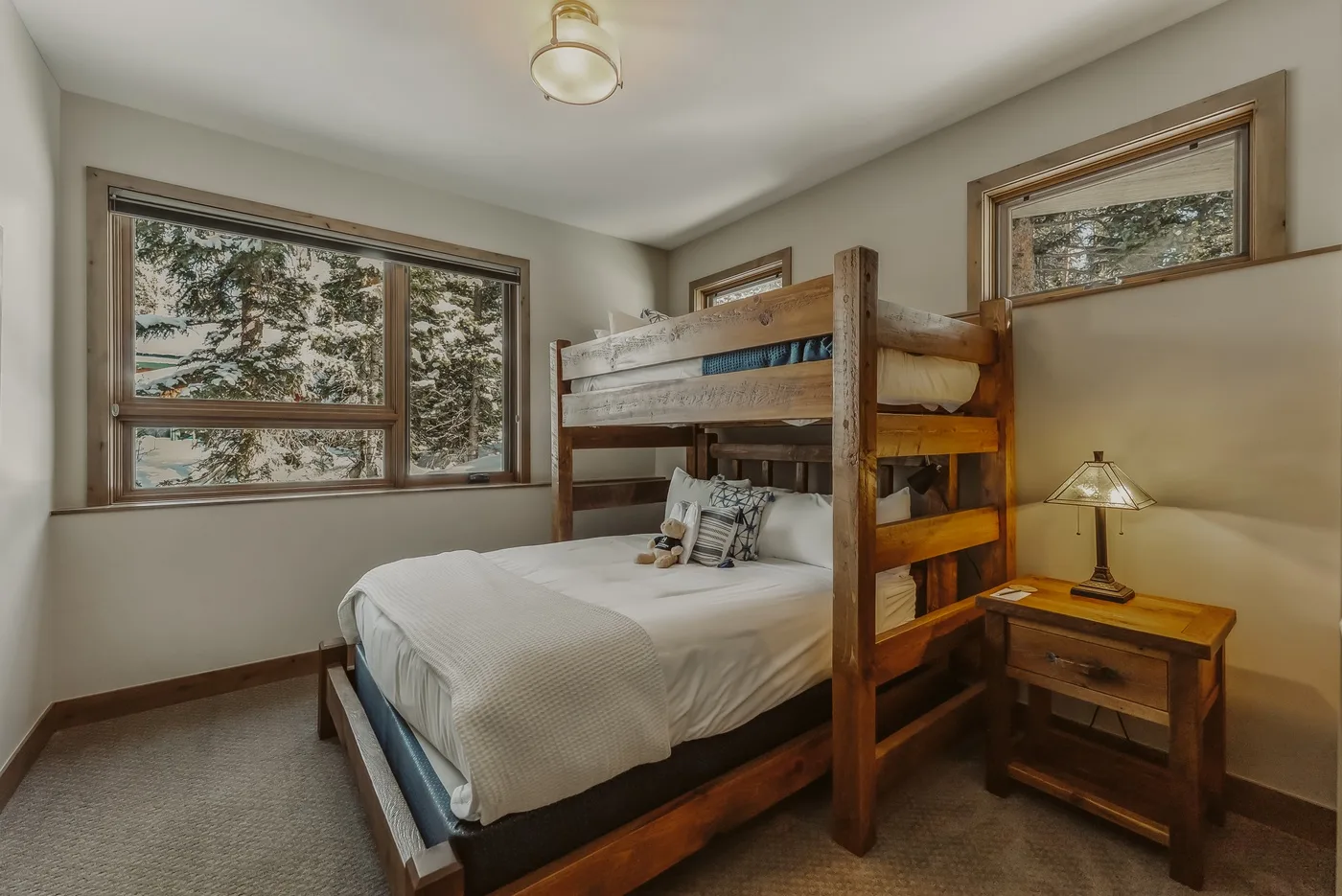 A bedroom with a wooden bunk bed, a lower bed, and windows overlooking trees.