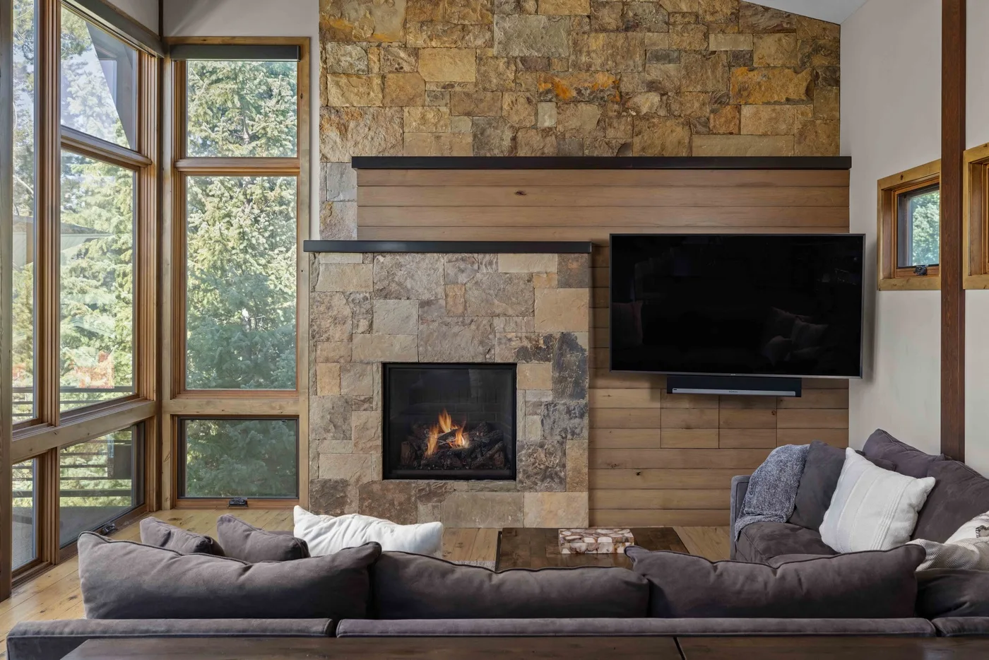 A living room with a stone fireplace, television, and seating beside tall windows.