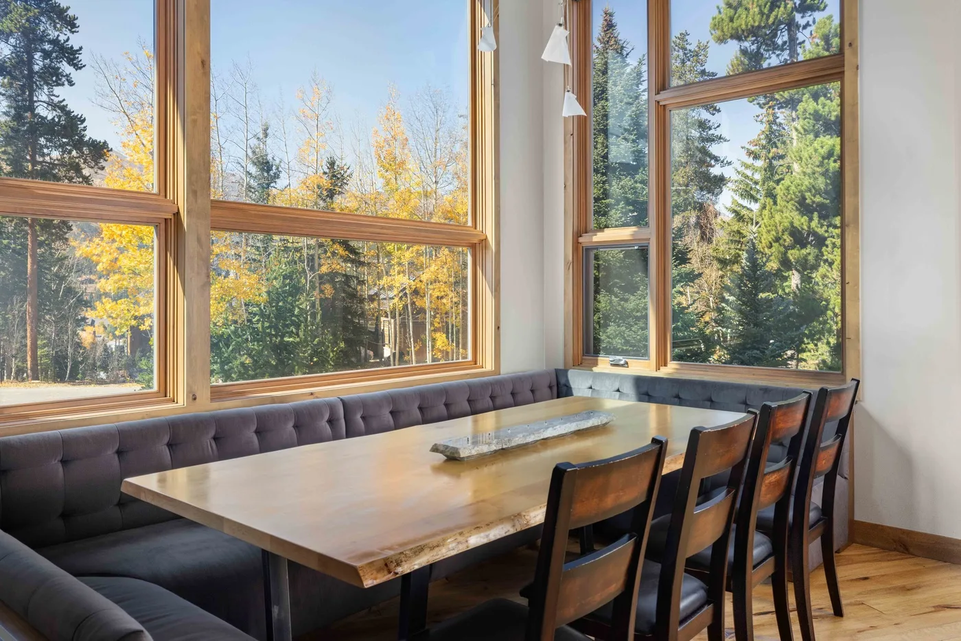 A dining area with a long table and seating beside large windows overlooking trees.