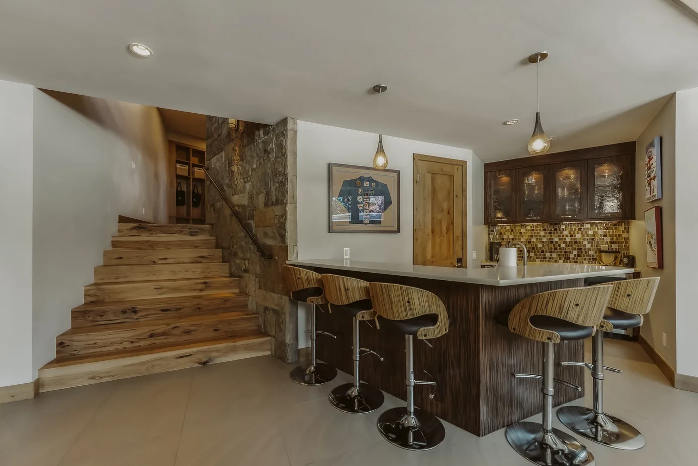 A bar counter with stools sits beside a wooden staircase in an indoor lounge.
