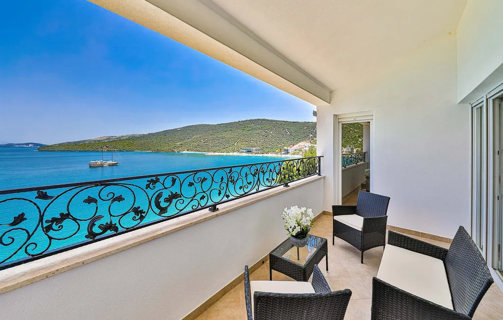 A balcony seating area overlooks the blue sea and a green coastline.