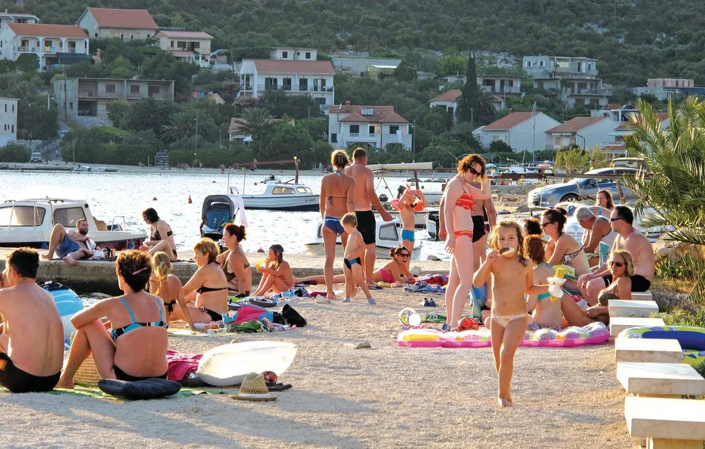Una playa concurrida con personas tomando el sol y nadando junto a embarcaciones.