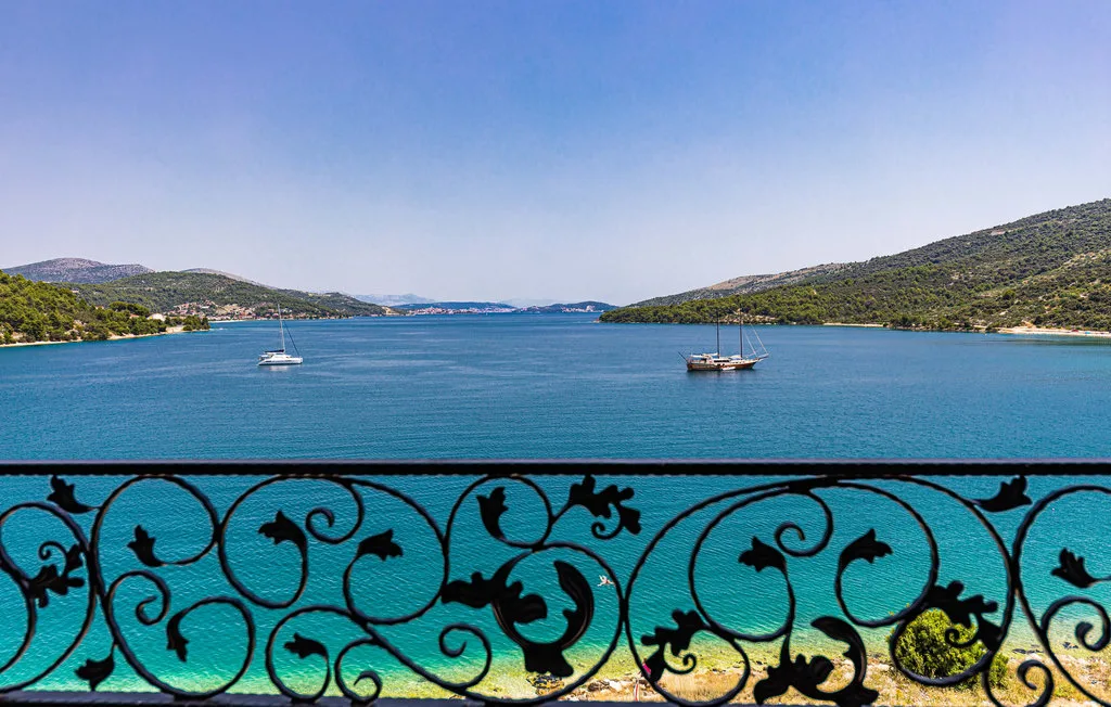 A blue bay with sailboats is viewed over an ornate black railing.