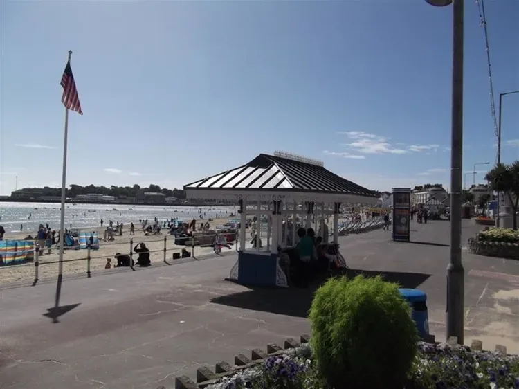 A seaside promenade with a small shaded kiosk, beach, and people along the shore.