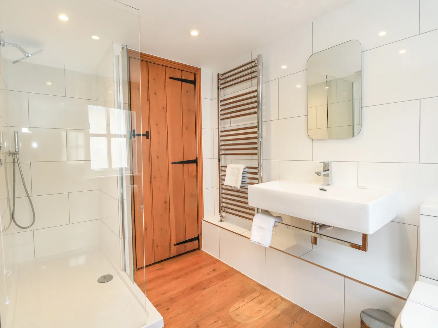A bright bathroom with a glass shower, wooden door, and wall-mounted sink.