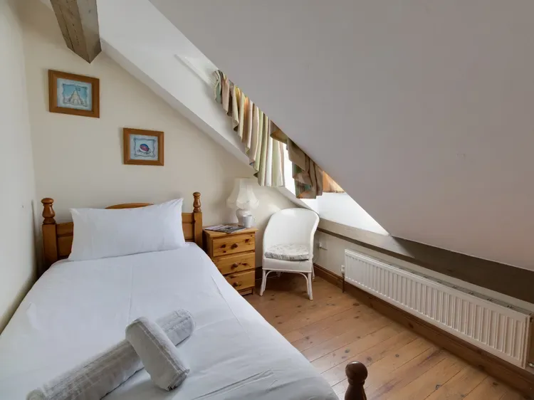 A bedroom with a white bed, wooden nightstand, and small window beneath a sloped ceiling.