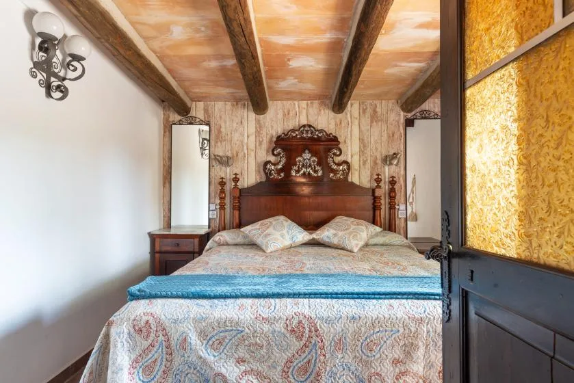 Un dormitorio con una cama de madera tallada y ropa de cama estampada bajo vigas vistas.