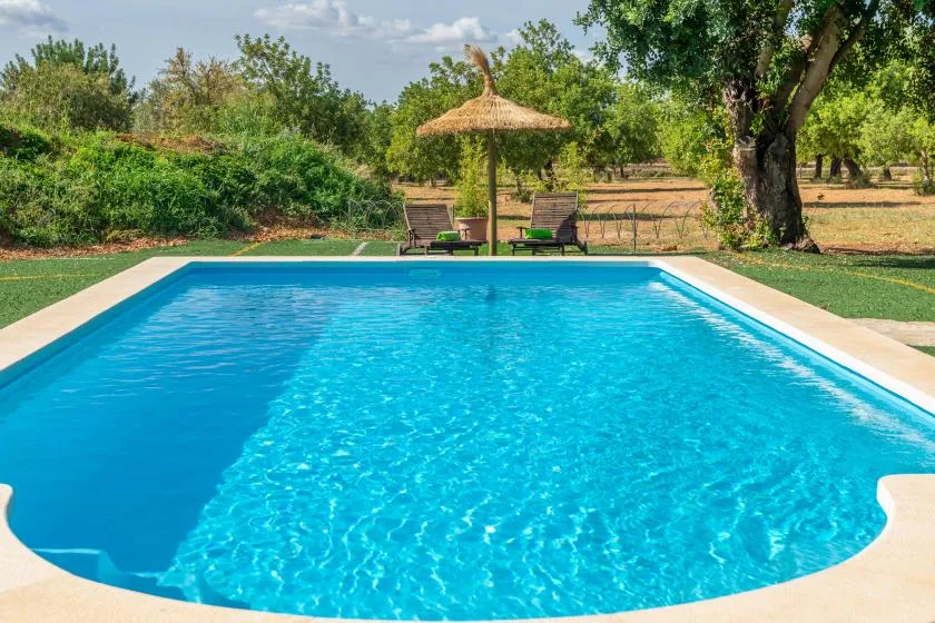 Una piscina al aire libre está junto a un jardín con césped y dos tumbonas bajo una sombrilla.
