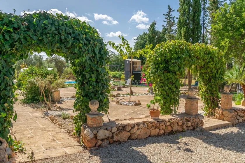 Un patio ajardinado con arcos cubiertos de enredaderas, caminos de piedra y árboles.