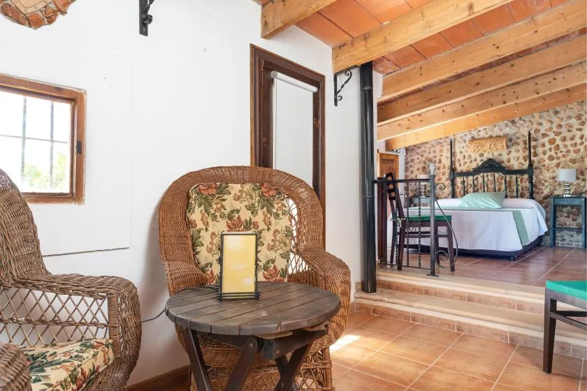 Dormitorio rústico con una cama visible detrás de una zona de estar con asientos de mimbre.