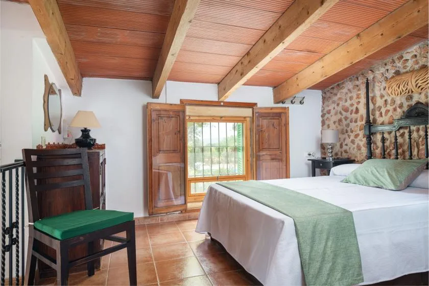 Un dormitorio con una cama, vigas de madera en el techo y una ventana entre contraventanas de madera.