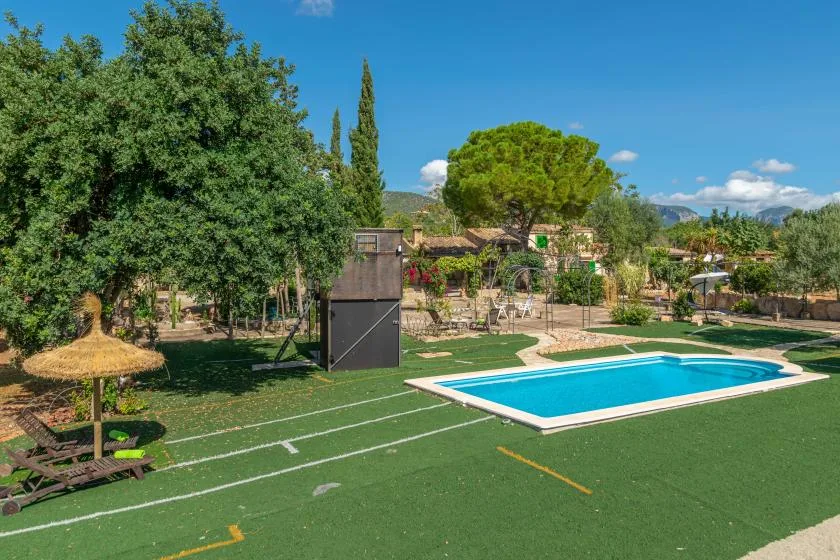 Una piscina al aire libre está junto a una cancha deportiva verde, con árboles y asientos cerca.