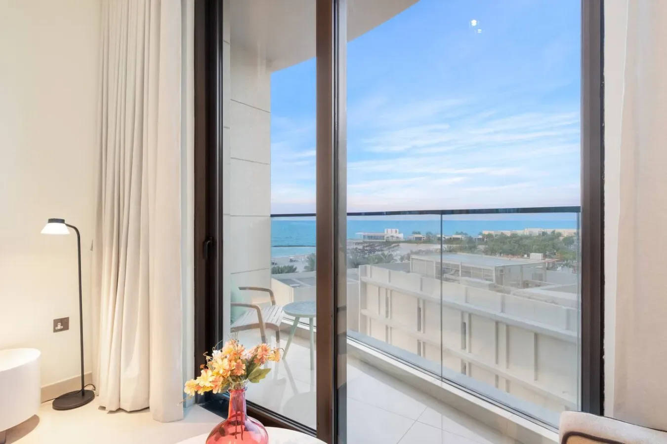 Una habitación luminosa da a un balcón con vistas al mar y a edificios cercanos.