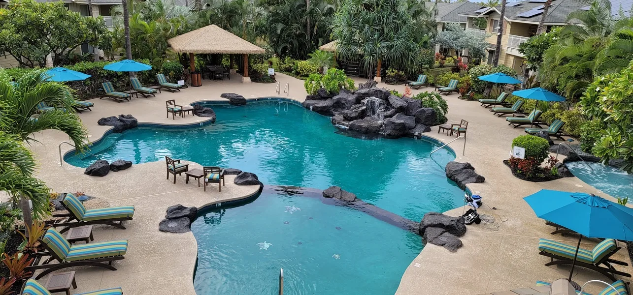 A resort-style outdoor swimming pool surrounded by seating, umbrellas, and tropical greenery.