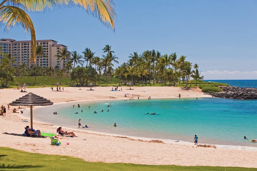 A sunny beach curves around turquoise water, with palm trees and resort buildings in the background.