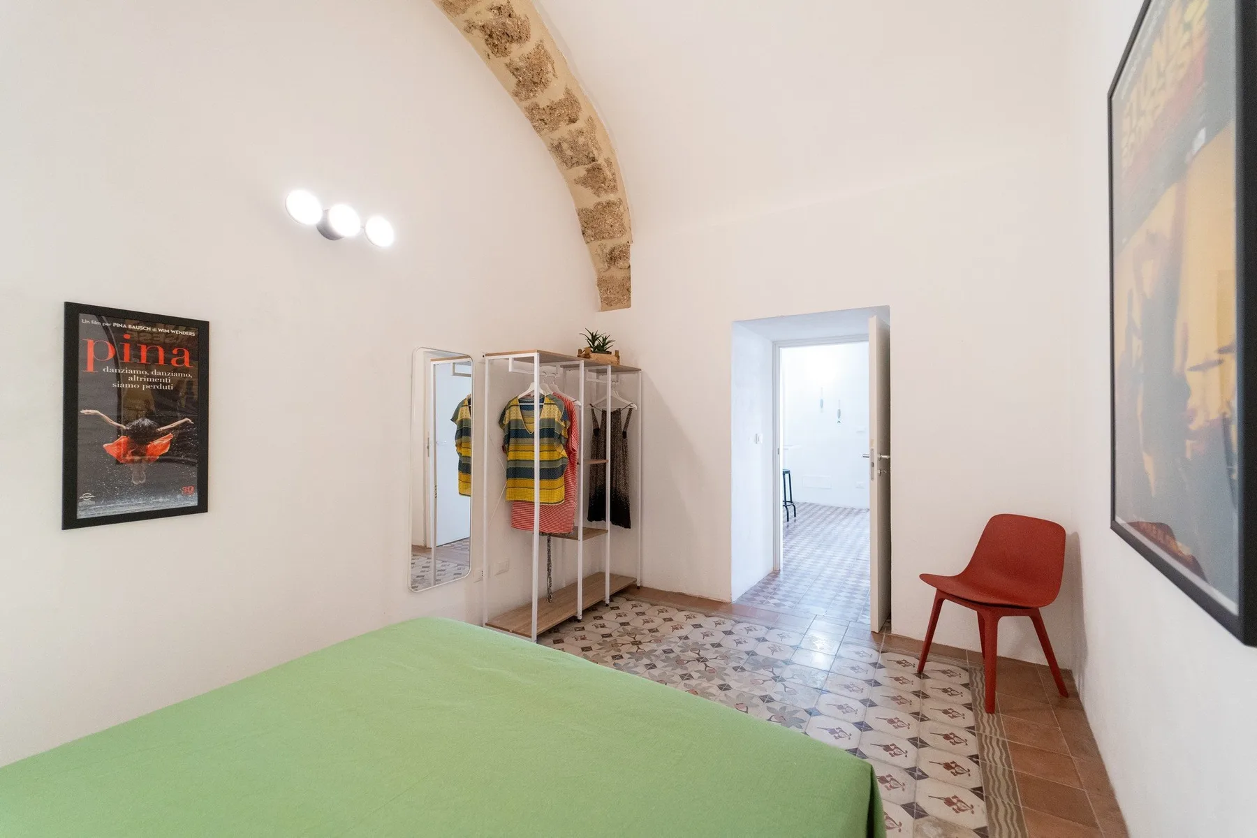 A bedroom with a green bedspread, clothing rack, and doorway to a tiled room.