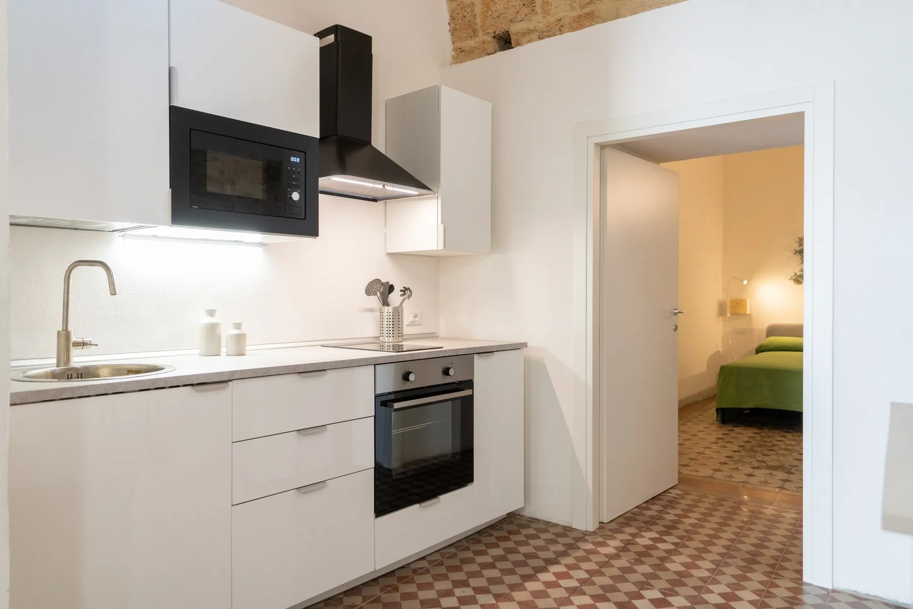 A compact kitchen opens through a doorway to a bedroom with green bedding.